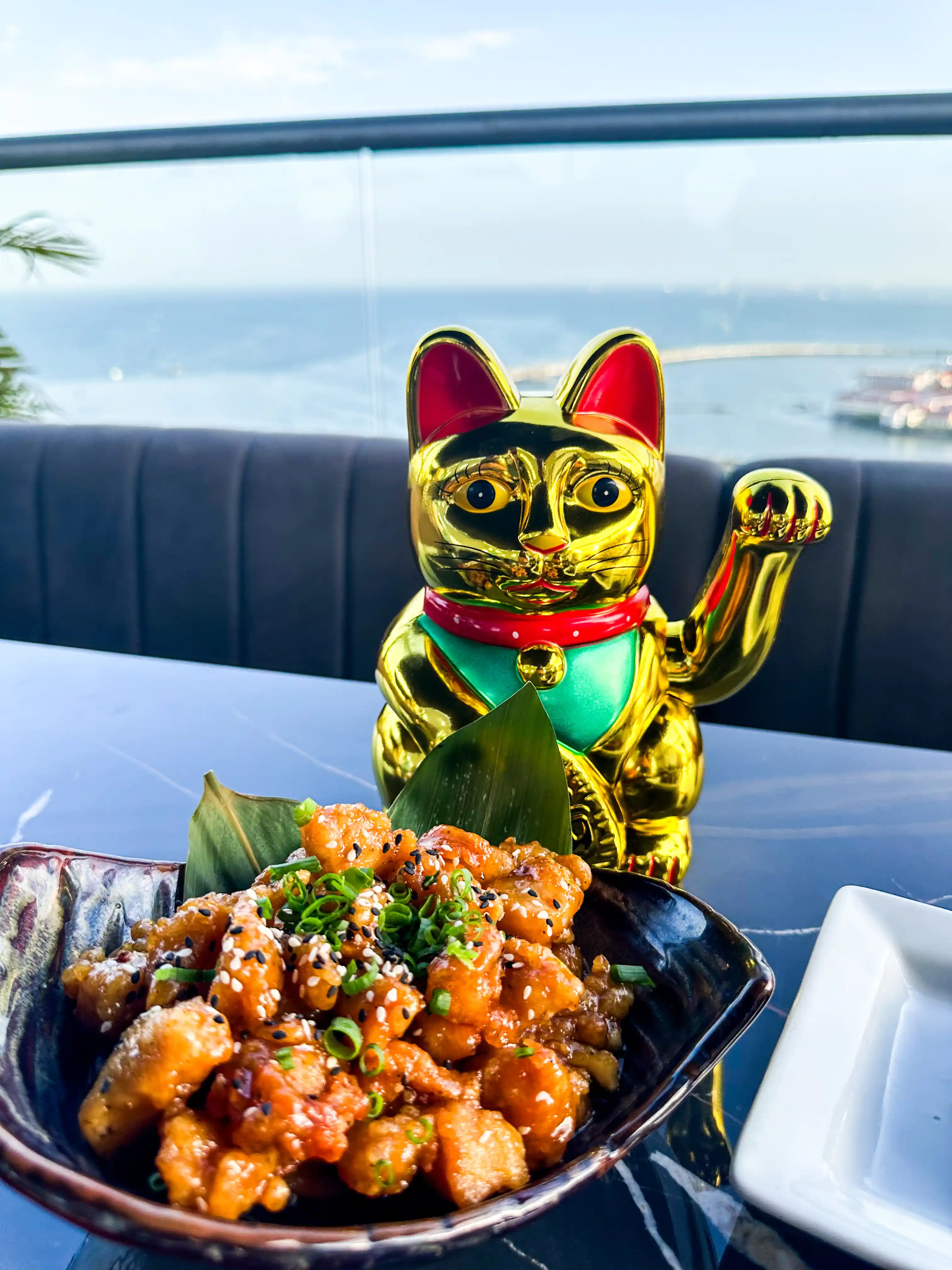 Una estatua de gato de la suerte dorada junto a un vibrante plato de pollo al sésamo, ambientado con una vista escénica al océano en un elegante restaurante en la azotea