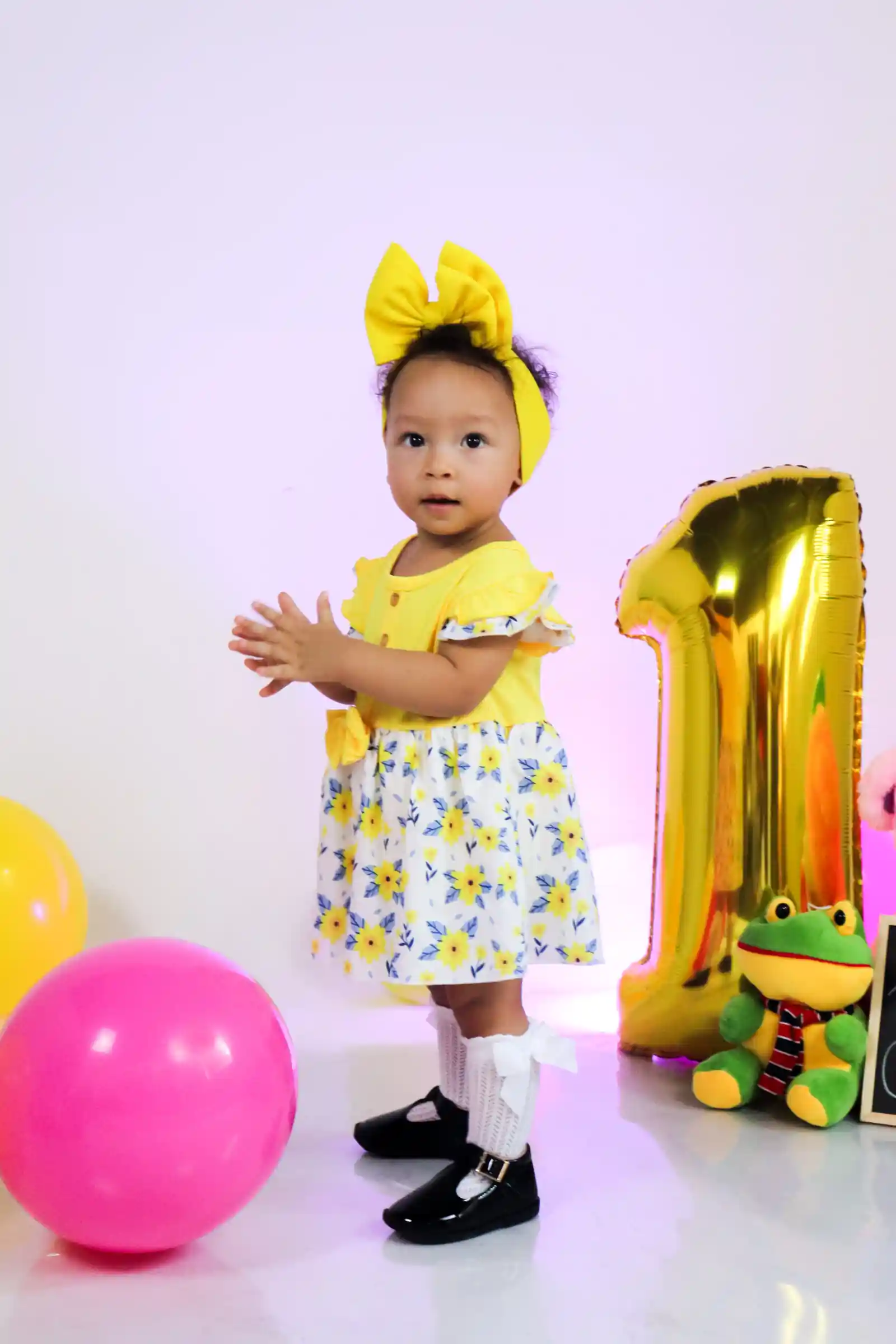 Un niño pequeño alegre en un vestido amarillo aplaude junto a globos y un juguete de rana juguetón en su primer cumpleaños